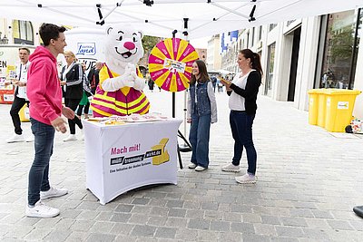 Person in Bärenkostüm mit gestreiftem T-Shirt steht hinter einem Tisch mit Aufschrift 'Mach mit! Müll trennen wirkt.de' neben einem Glücksrad auf einem Bürgersteig