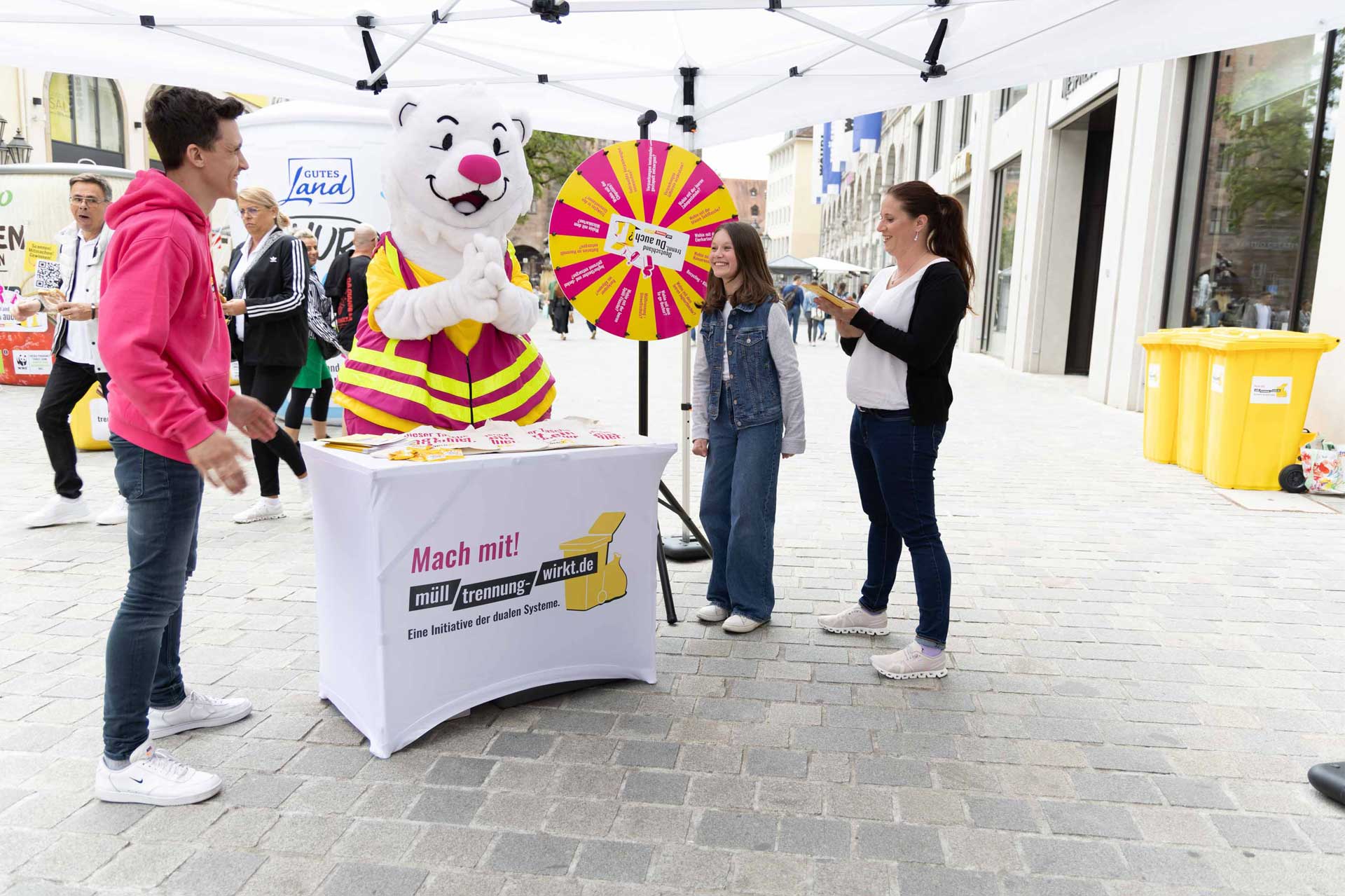 Person in Bärenkostüm mit gestreiftem T-Shirt steht hinter einem Tisch mit Aufschrift 'Mach mit! Müll trennen wirkt.de' neben einem Glücksrad auf einem Bürgersteig